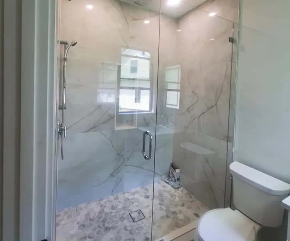 Walk-in shower with marble-look tile walls, a hexagon mosaic floor and a hinged glass door, next to a toilet