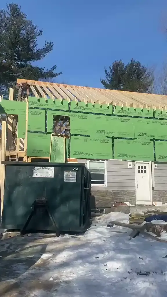 Home addition walls in green ZIP System sheathing with open roof rafters above a gray-sided first floor, with a dumpster in front, in Lynnfield, MA (video still)