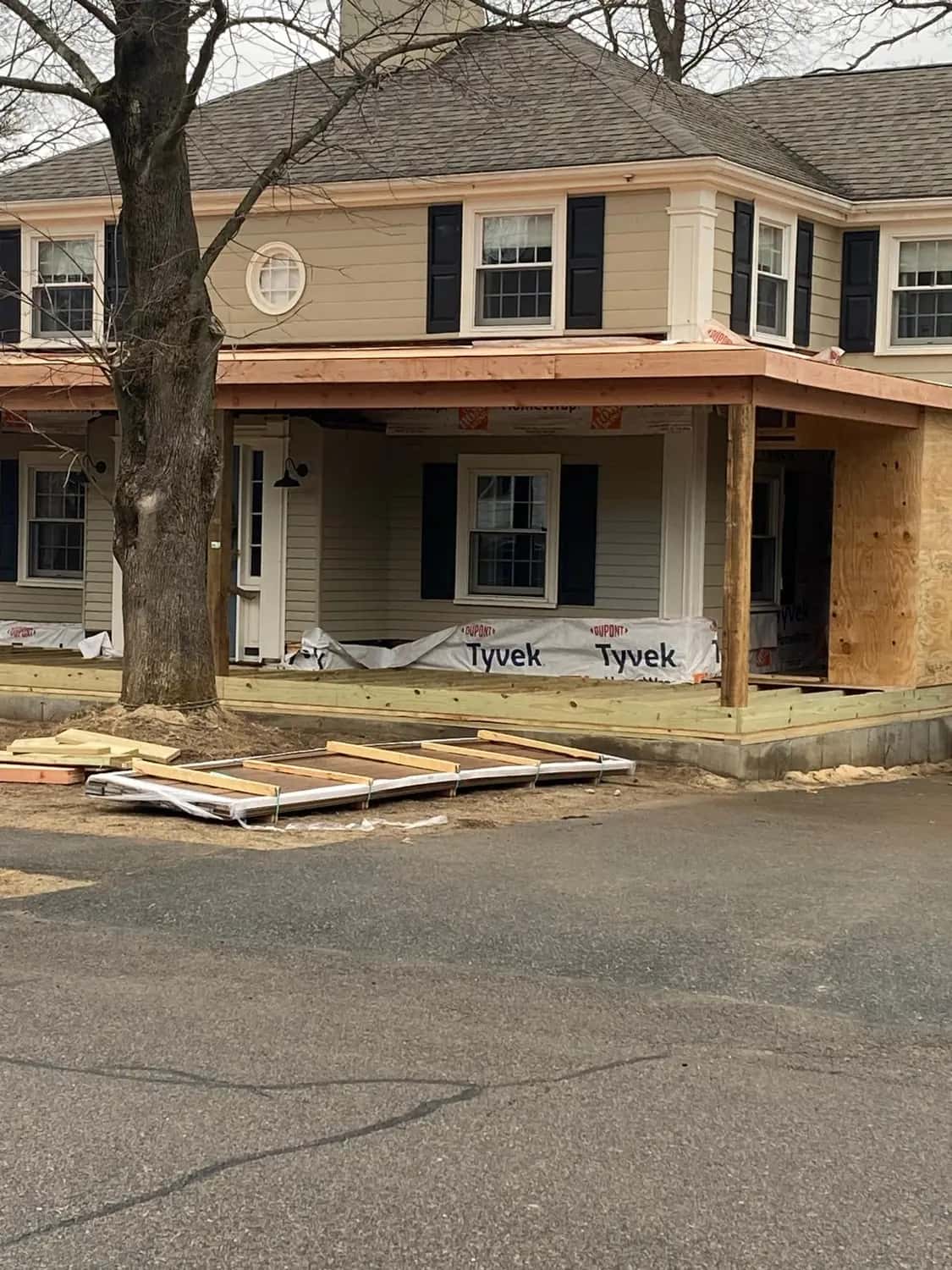Colonial house with a new front porch roof frame, house wrap along the base and a plywood-sheathed section at the side (Needham, MA)